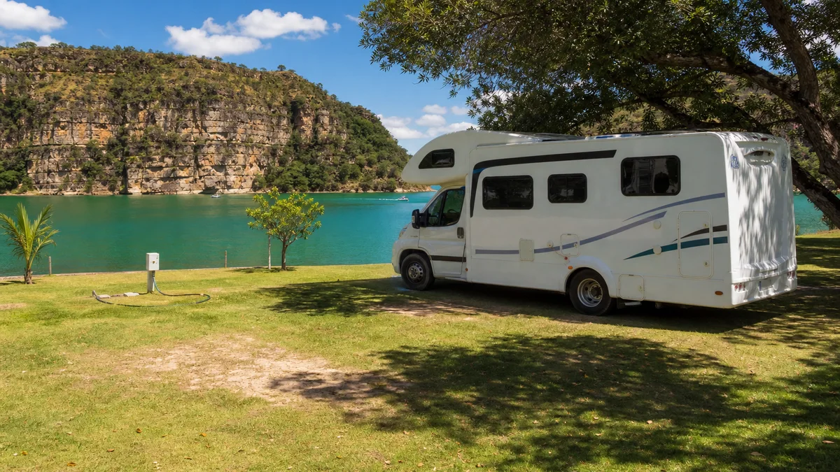 Motorhome em camping à beira de lago verde com paredões ao longe ao sol da tarde