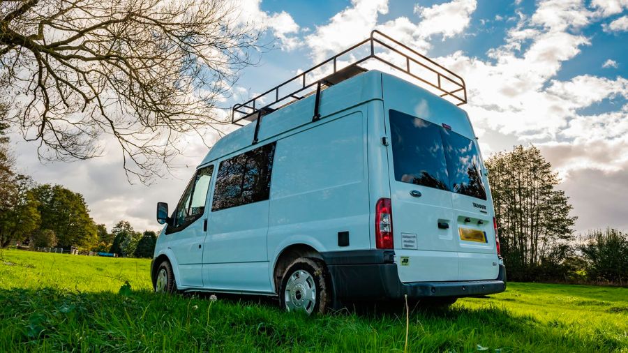 Van adaptada branca estacionada em área verde com céu parcialmente nublado, usada como imagem ilustrativa no artigo